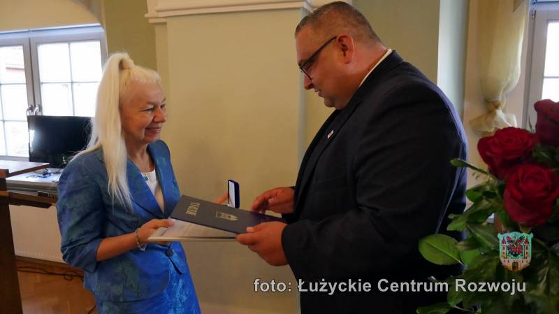 mężczyzna w ciemnej marynarce wręcza medal i teczkę z dokumentami kobiecie w niebieskim żakiecie