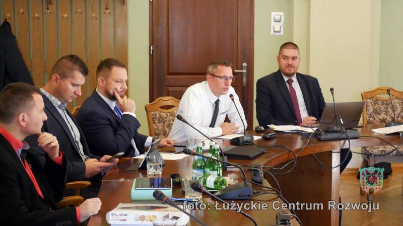 pięciu mężczyzn siedzi przy stole, mężczyzna w białej koszuli mówi do mikrofonu stojącego przed nim