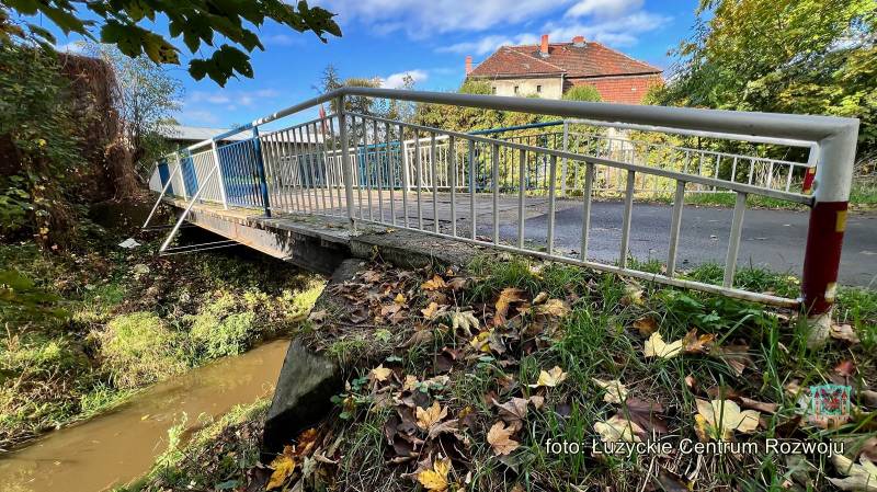 Widok na barierkę od mostu, na pierwsym planie trawa z jesiennymi liśćmi. Pod mostem płynie rzeka, w tle budynek.